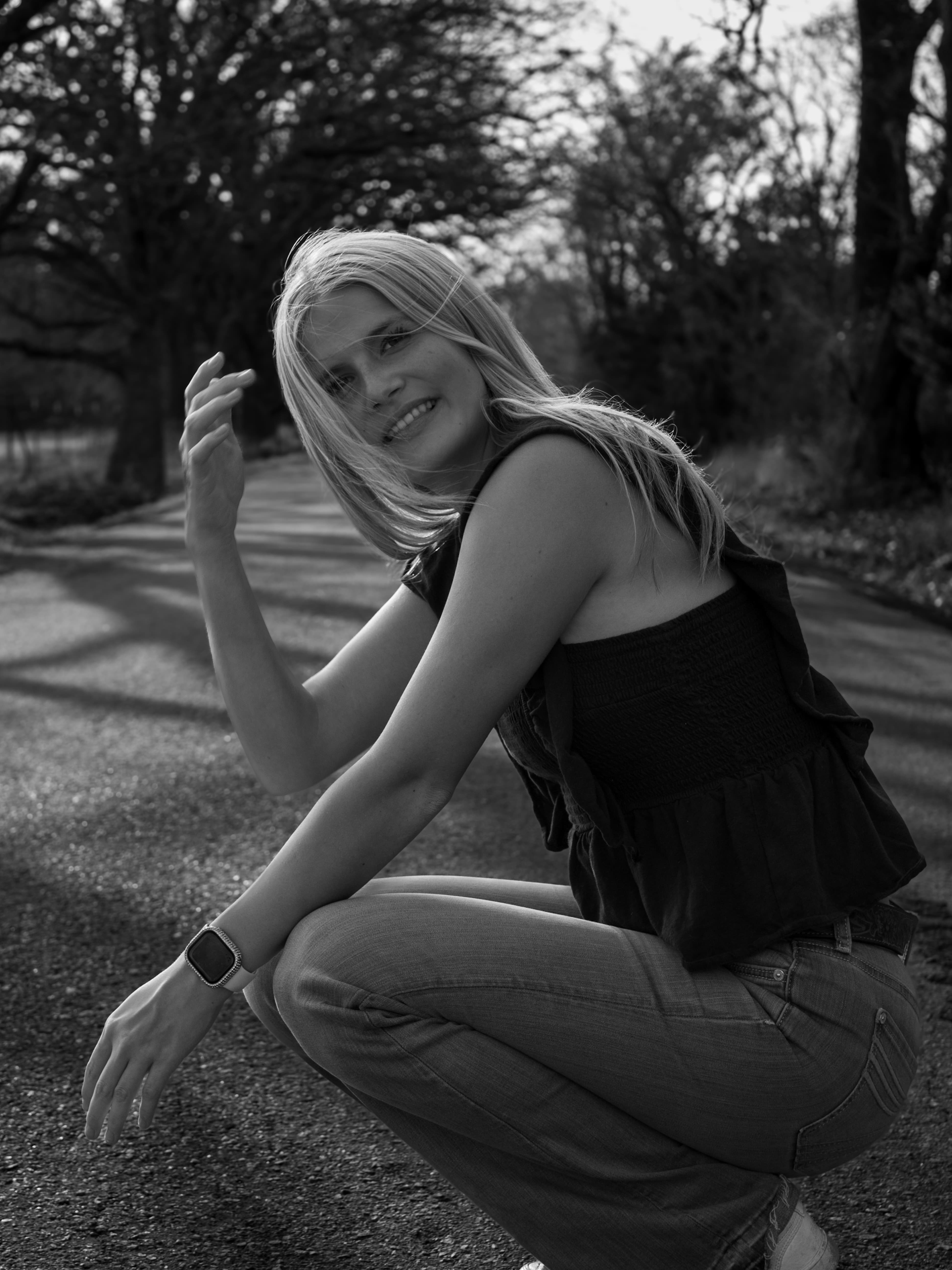 A black-and-white portrait of a smiling woman crouched on a tree-lined road.