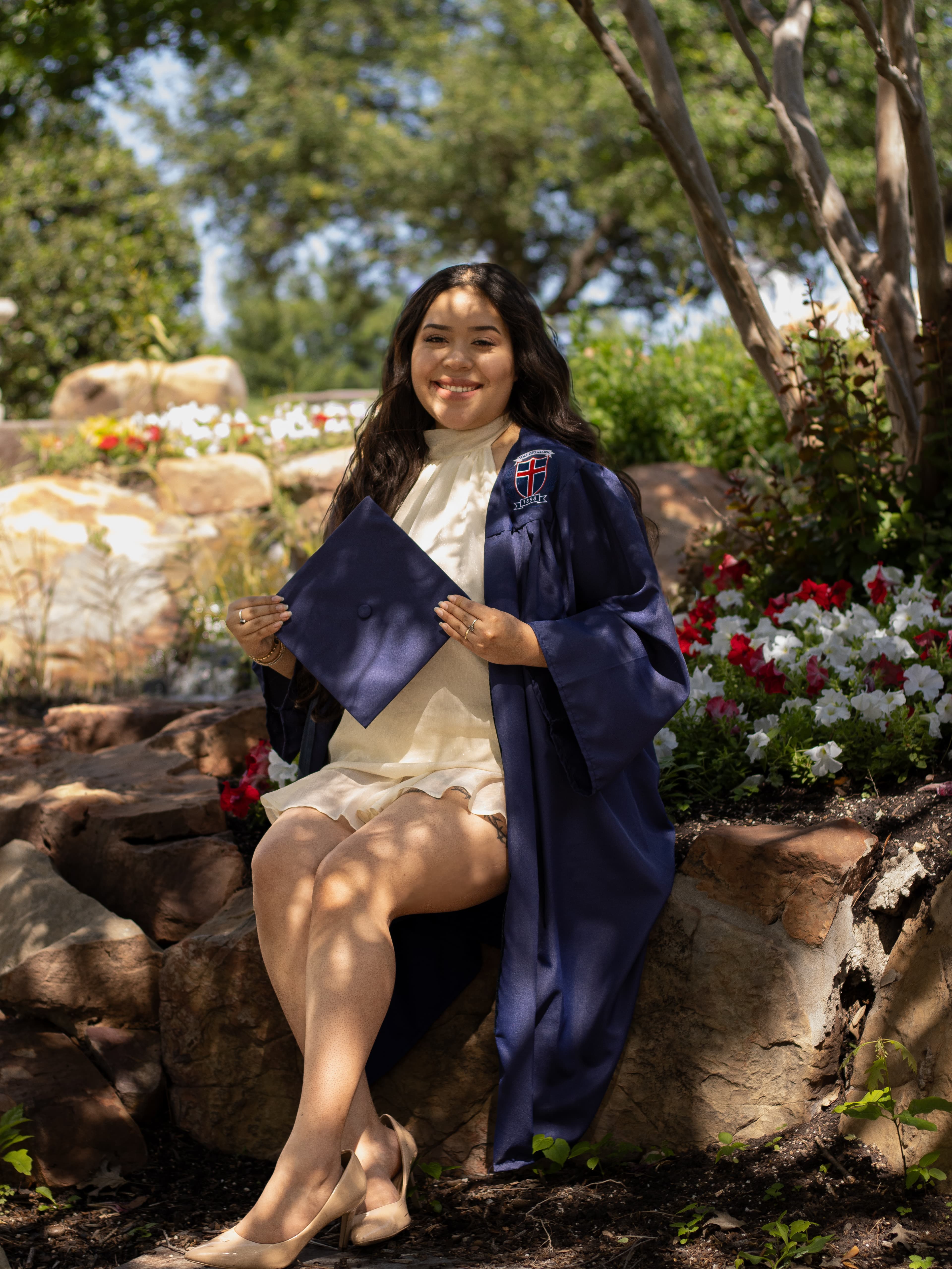 A graduate in a navy gown seated among garden rocks and flowers.