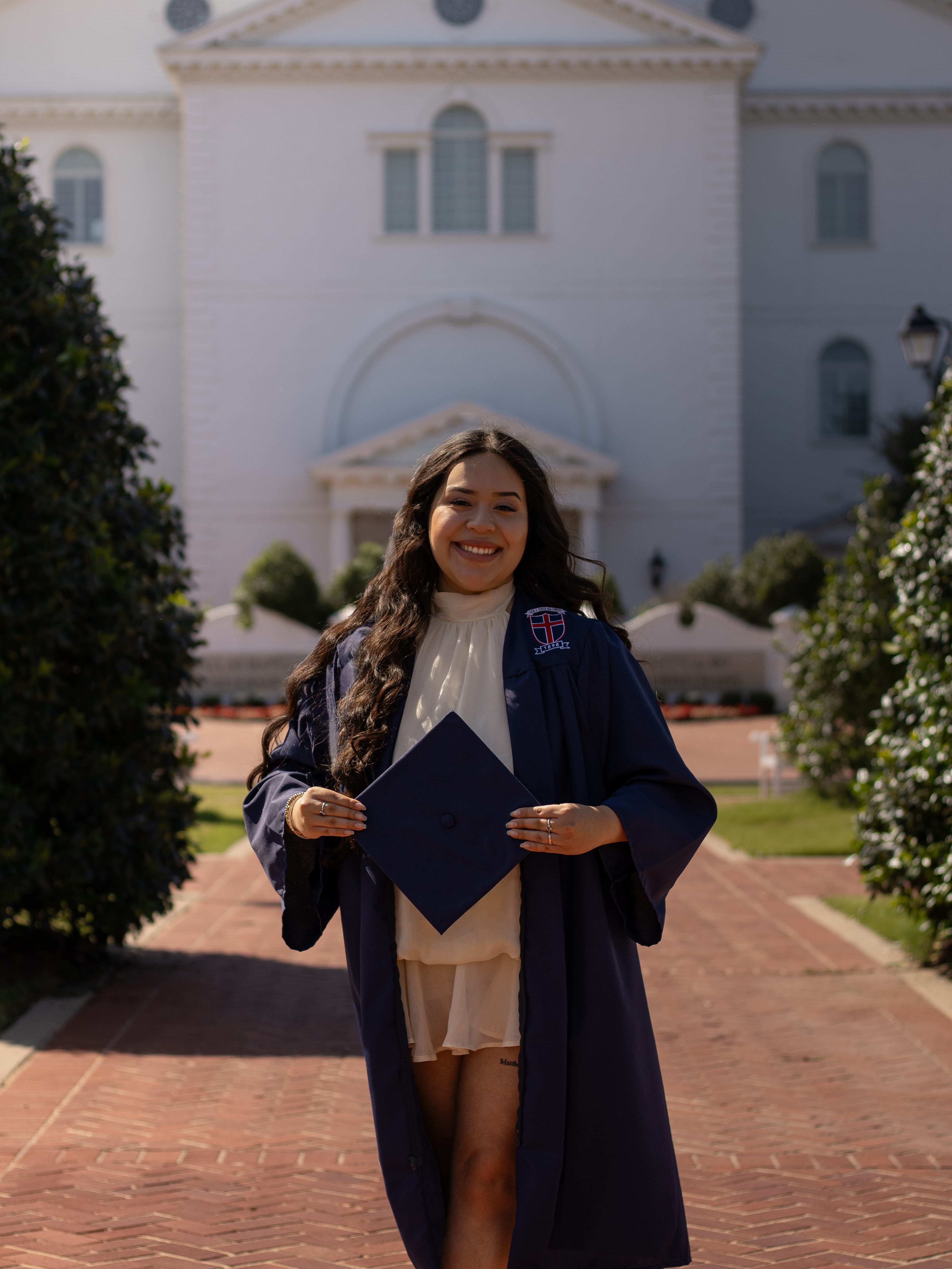 A smiling graduate holding her cap on a brick campus walkway.
