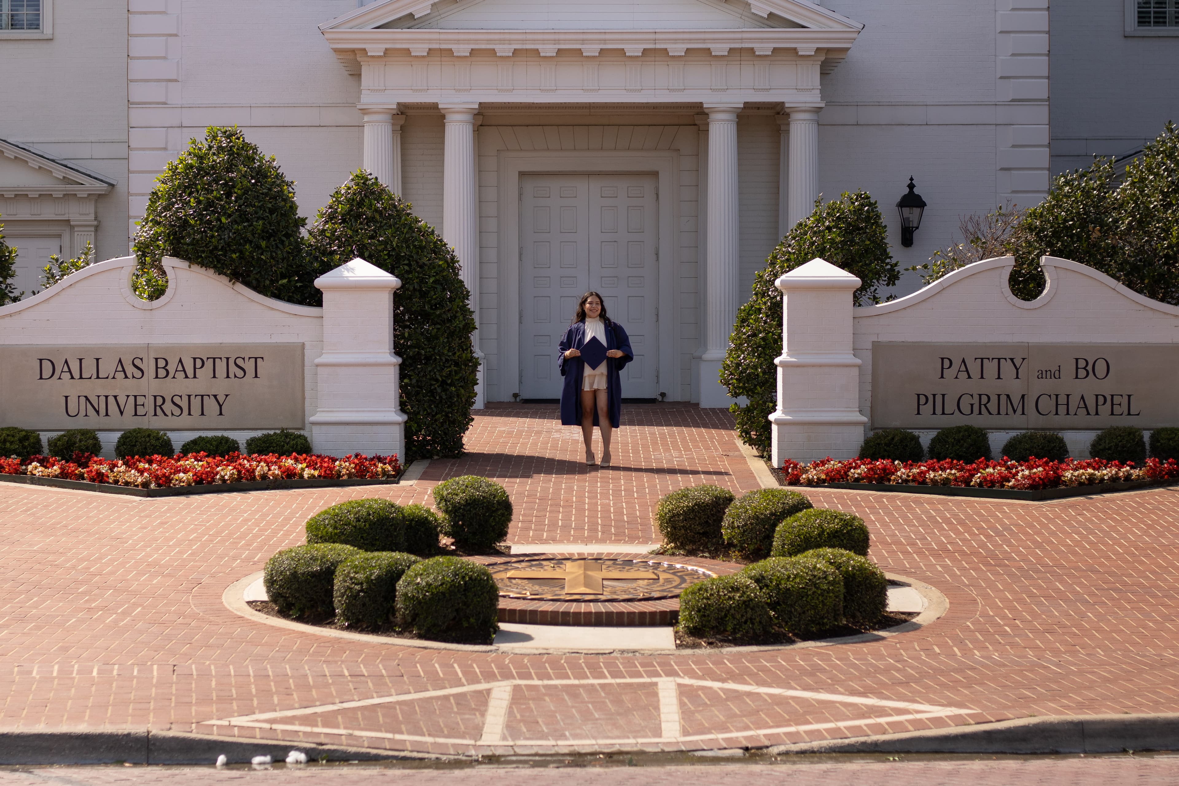 A graduate standing at the Dallas Baptist University entrance, framed by flowers and white columns.