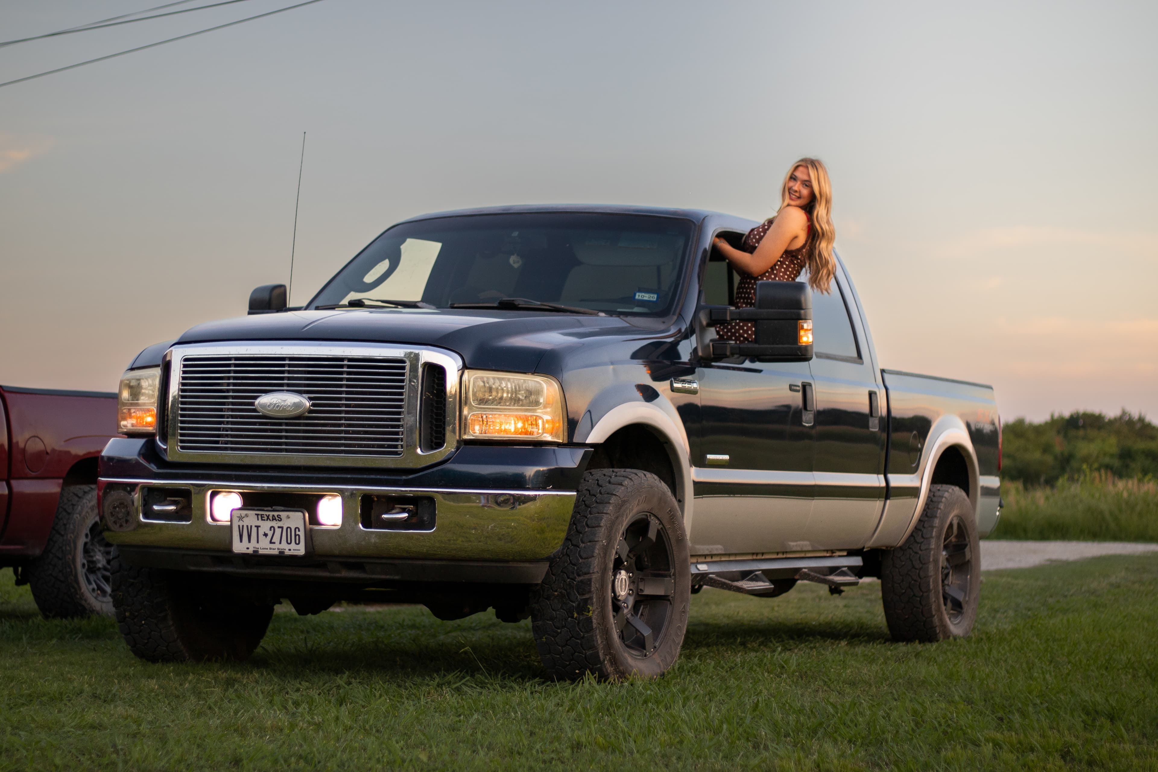 A woman sitting on a pickup truck in a grassy field at dusk.
